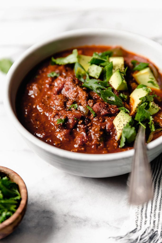 A white bowl filled with thick and hearty pumpkin chili topped with diced avocado and sliced scallions.