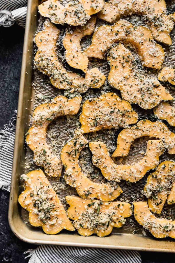 Acorn squash slices coated in Parmesan cheese and fresh herbs on a sheet pan ready for roasting.
