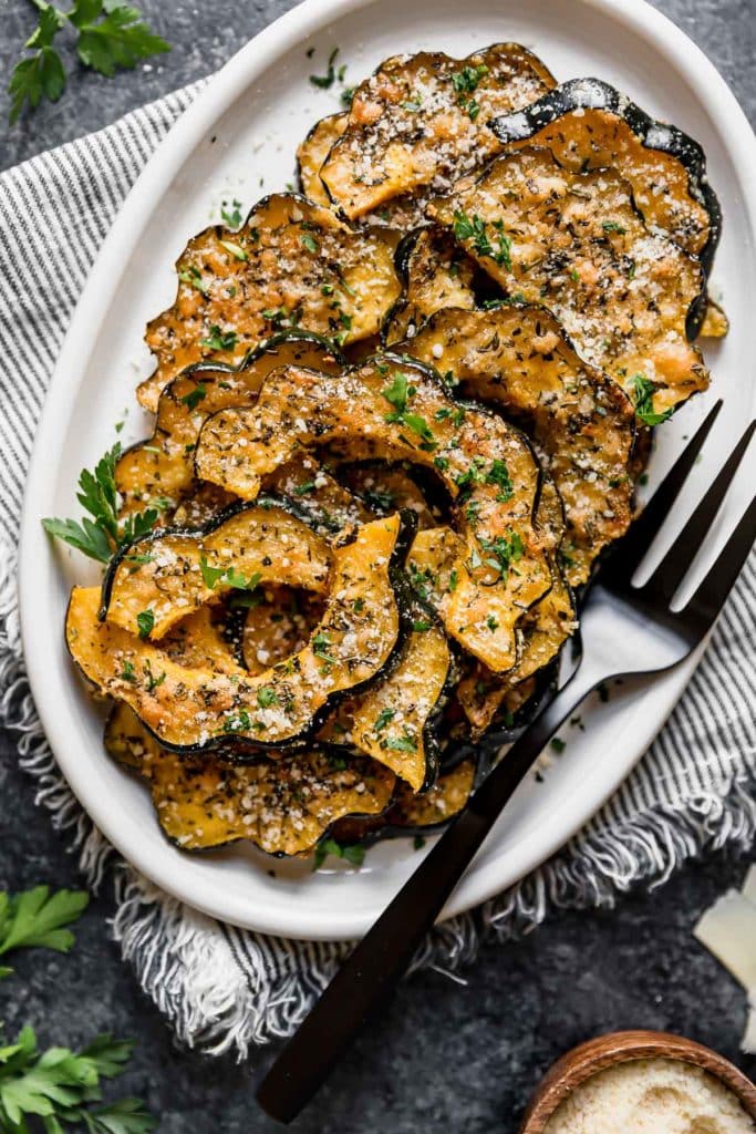 White platter filled with acorn squash slices baked with parmesan cheese and fresh herbs, black serving fork on side.