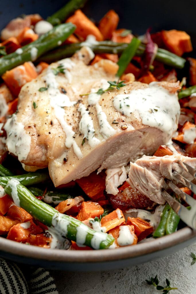 Close up view of a cut piece of chicken with vegetables surrounding, topped with ranch sauce and fresh herbs. 