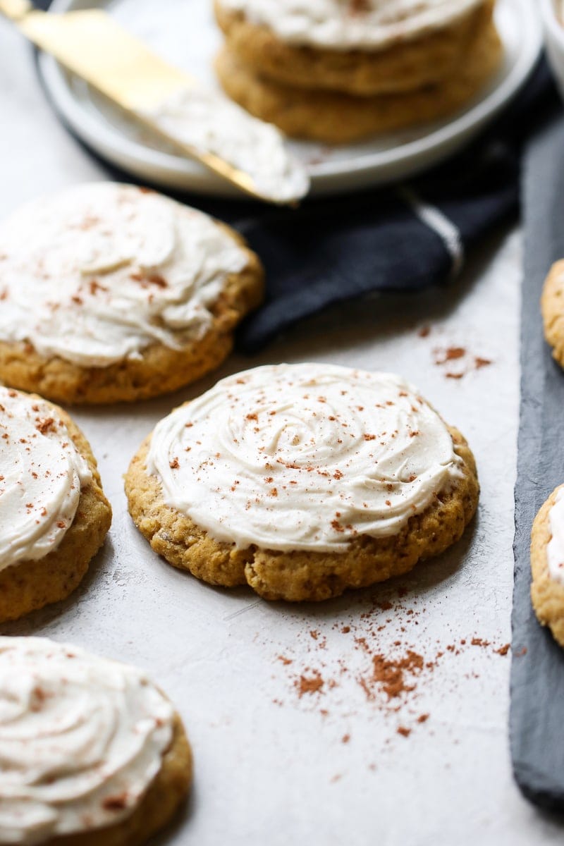 Several Soft Pumpkin Cookies with Cream Cheese Frosting, dusted with pumpkin spice on a white table.