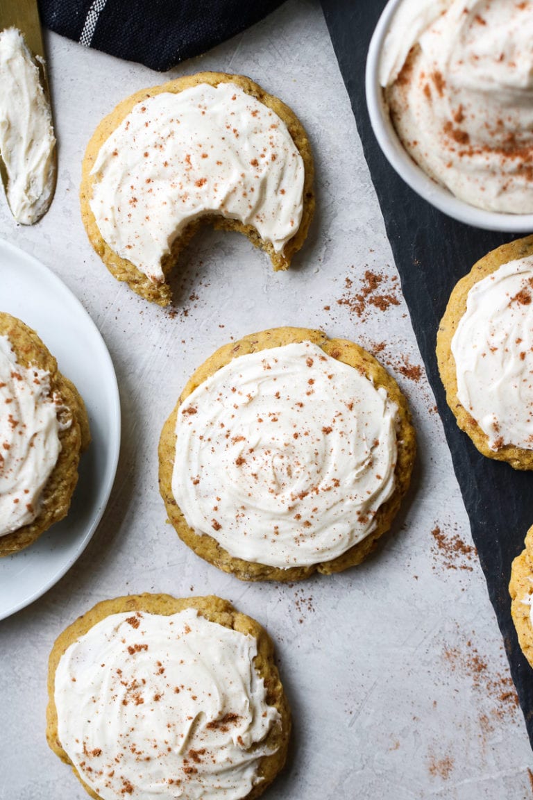 Soft pumpkin cookies with cream cheese frosting on a sheet of parchment paper, one cookie missing a bite.