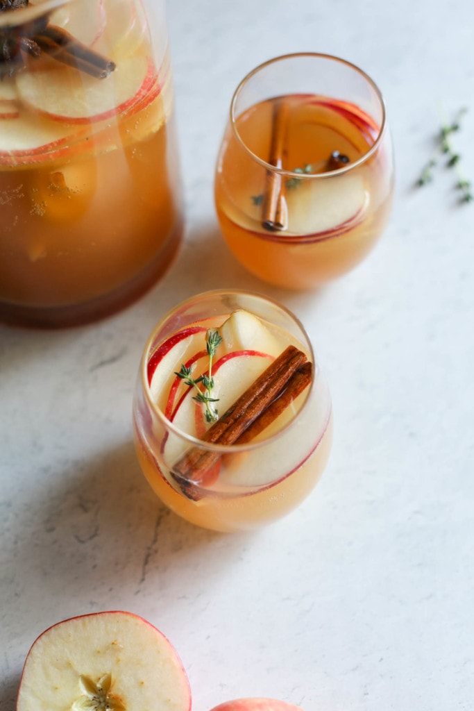 Two short glasses filled with apple cider sangria with cinnamon sticks and fresh thyme sprigs next to a pitcher of sangria.