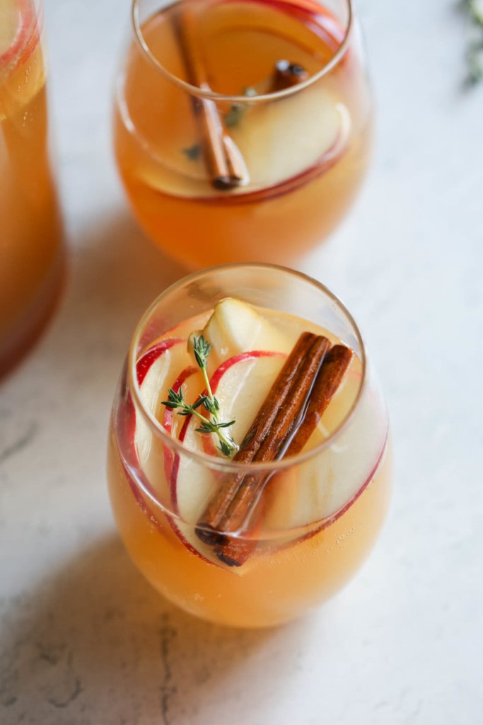 Close up view of apple cider sangria in a glass with apple slices, cinnamon sticks, and sprig of thyme floating in the glass.