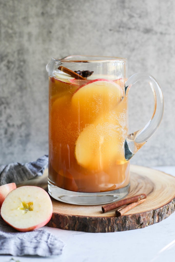 A tall glass pitcher filled with apple cider sangria with apple rounds, cinnamon sticks, and whole star anise floating in the sangria. 