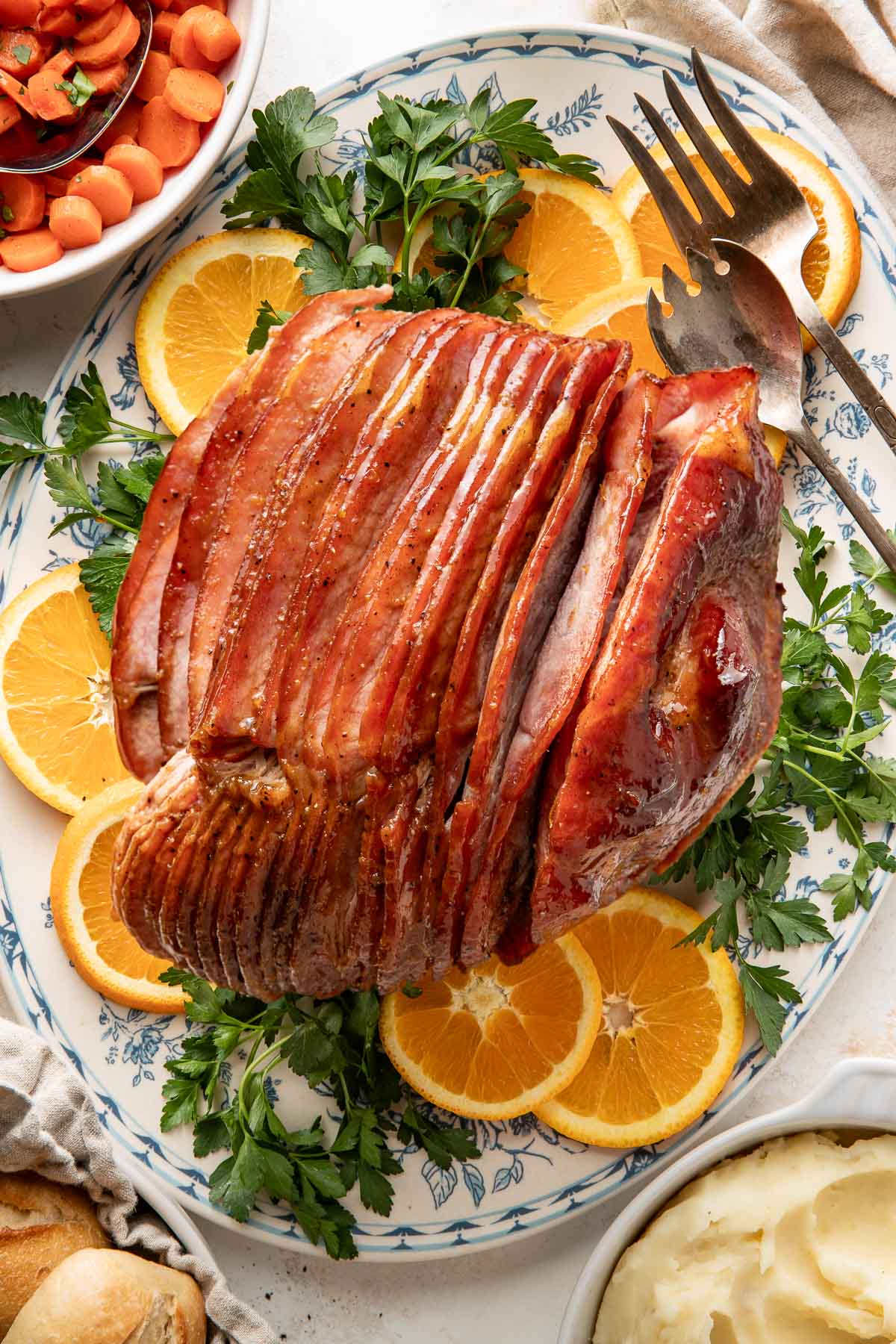 Overhead view of a sliced glazed ham on a platter surrounded by sliced oranges and fresh herbs. 