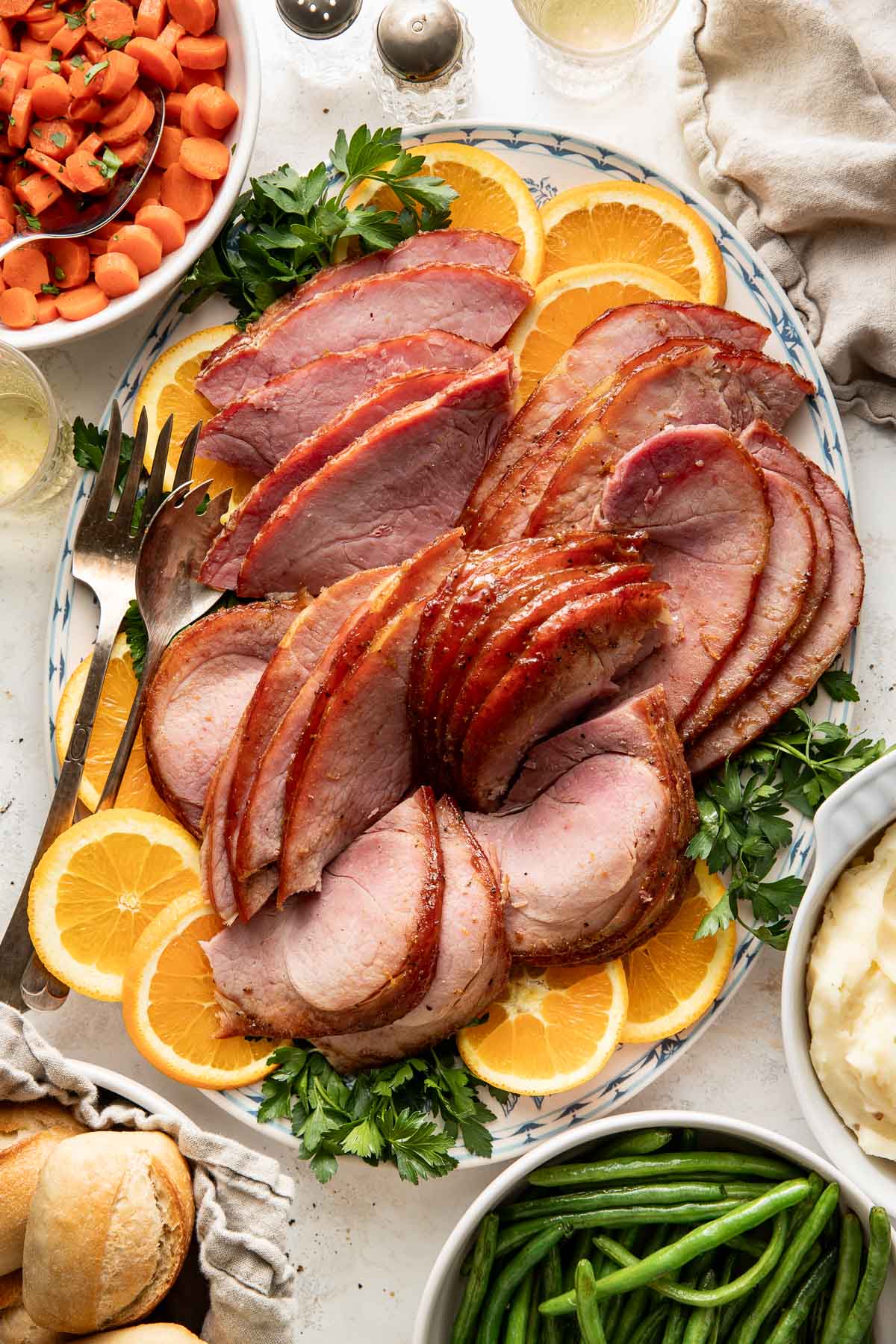 Overhead view of a platter of Baked Glazed Ham sliced and surrounded by oranges and fresh herbs.