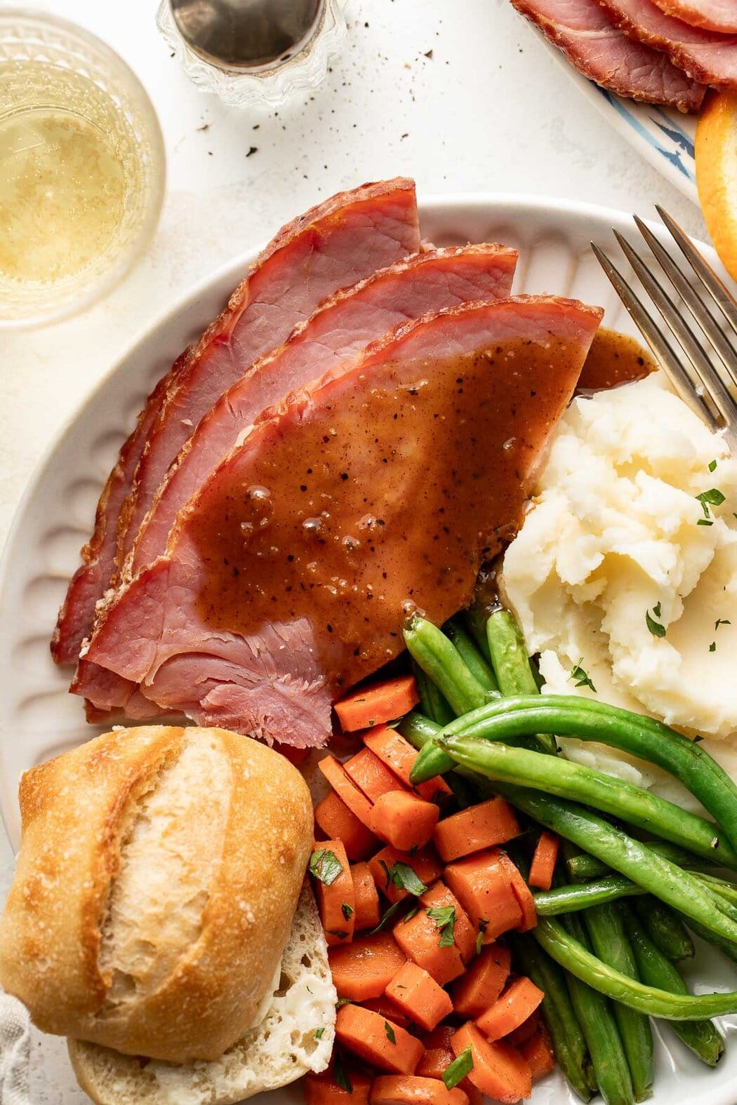 Overhead view of a plate filled with Baked Glazed Ham, mashed potatoes, green beans, candied carrots, and a dinner roll.