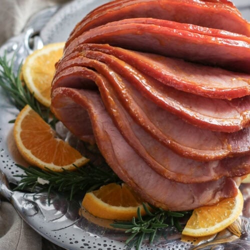 Slices of orange glazed ham fall onto one another on a metal serving tray with a rosemary and orange garnish.