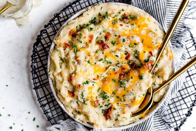 A white bowl filled with loaded mashed potatoes topped with melted cheddar cheese, chives, and bacon, ranch drizzled on top.