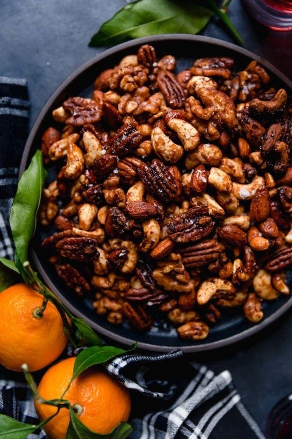 Overhead view of a black bowl on a dark grey surface filled with Slow Cooker Spiced Nuts (includes cashews, almonds, and pecans) with stem-on tangerines and a black and white plaid napkin lying next to the bowl. 