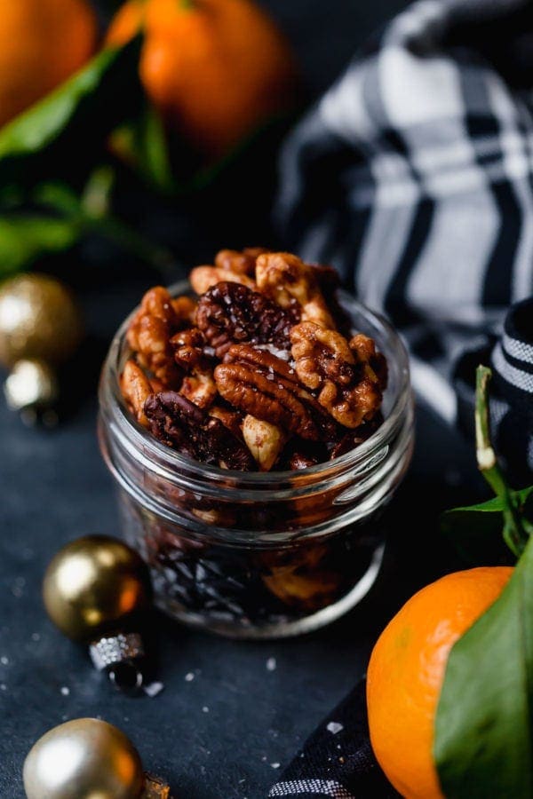 A small jar of Slow Cooker Spiced Nuts (includes cashews, almonds, and pecans) on a dark surface with tangerines, a black and white plaid napkin, and small gold ornaments surrounding the jar. 