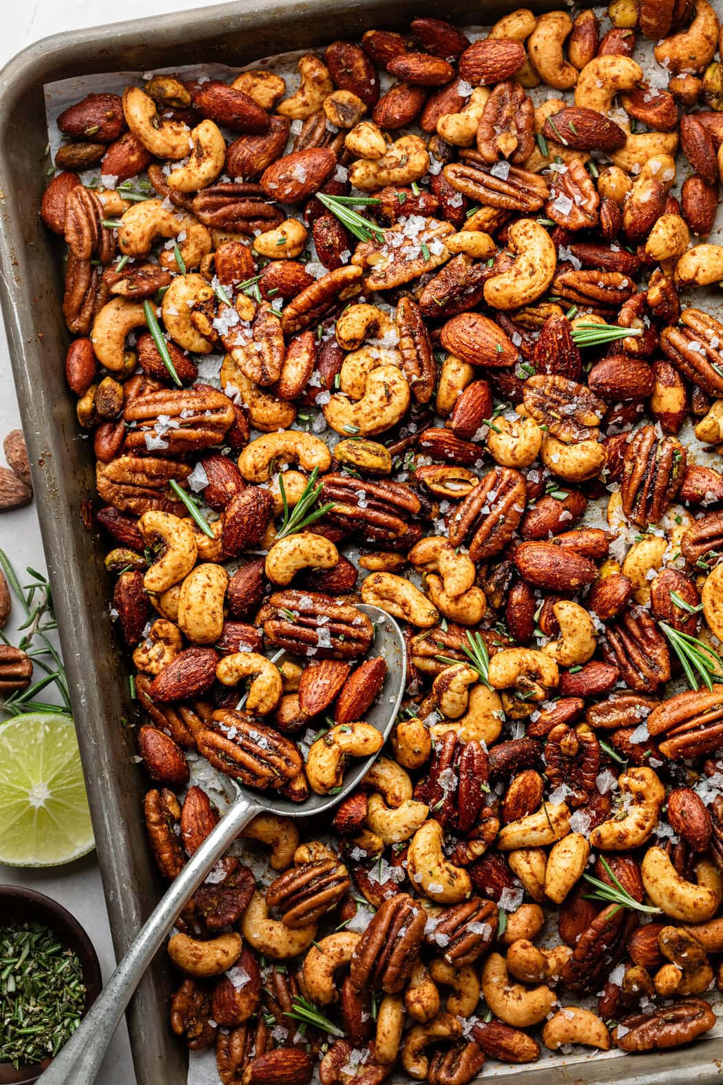 Chili rosemary roasted nuts on a baking sheet, a spoon lifting a scoop.