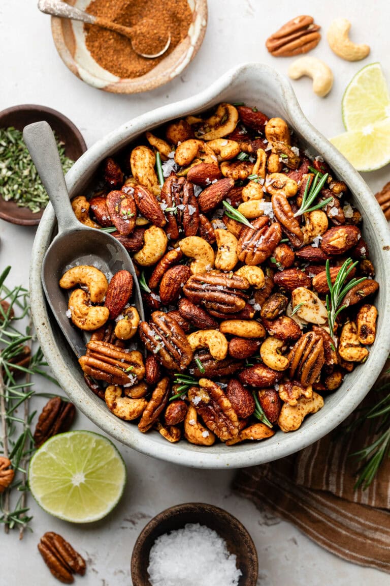 Close up view of a bowl of chili rosemary roasted nuts topped with flakey sea salt and fresh rosemary.