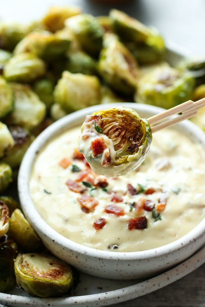 A roasted Brussels sprout half on a toothpick coated halfway with aioli sauce and bacon crumbles.