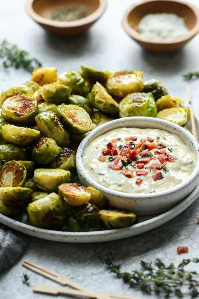 Close up view of roasted Brussels sprouts with a small bowl of aioli topped with bacon crumbles. 