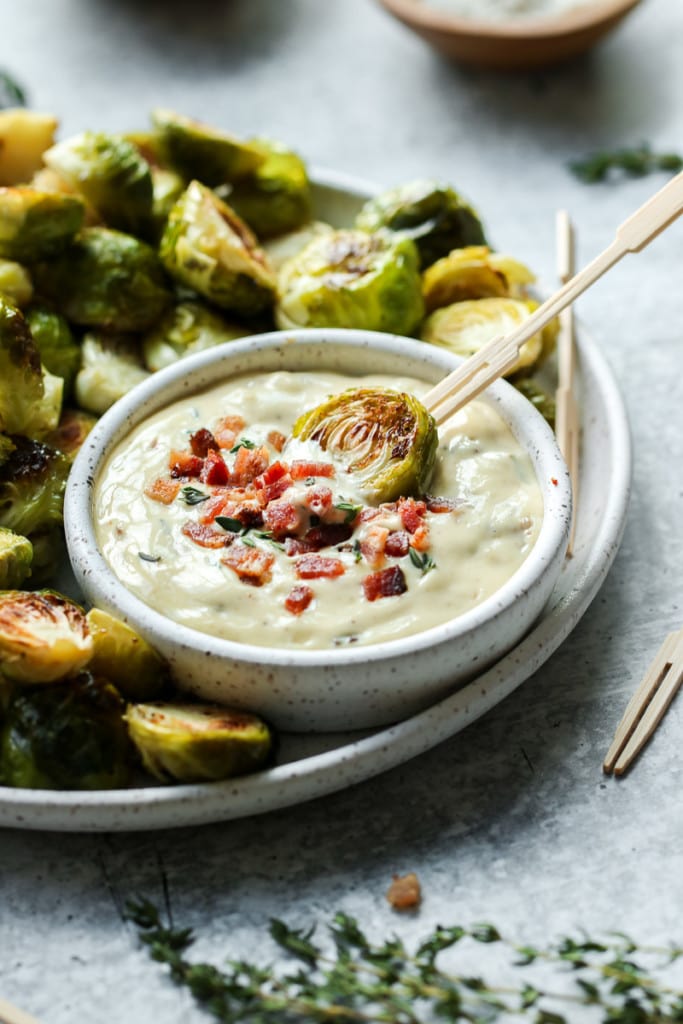 Close up view of a roasted brussels sprout half being dipped into aioli with bacon crumbles.