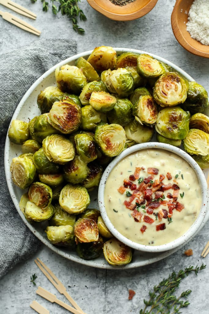 Overhead view of roasted Brussels sprouts on a speckled round platter with a small bowl of aioli sauce topped with chopped bacon.