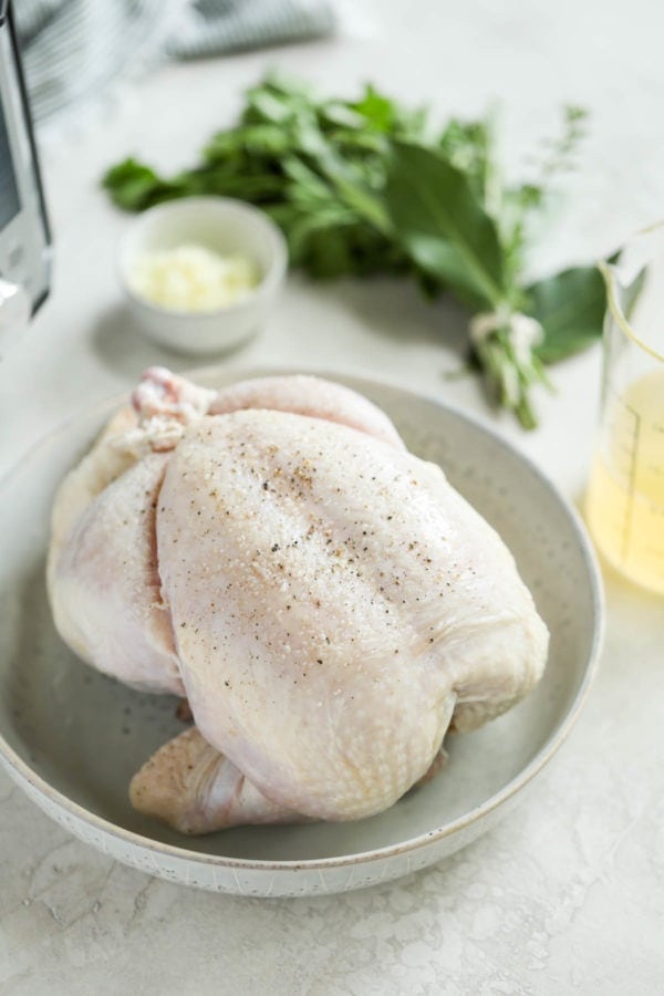 A raw, whole chicken in a bowl with fresh herbs, garlic and broth on the table next to it in preparation to make Instant Pot Whole Chicken. 