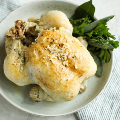 A roasted chicken in a cream speckled bowl with fresh herbs on the side.