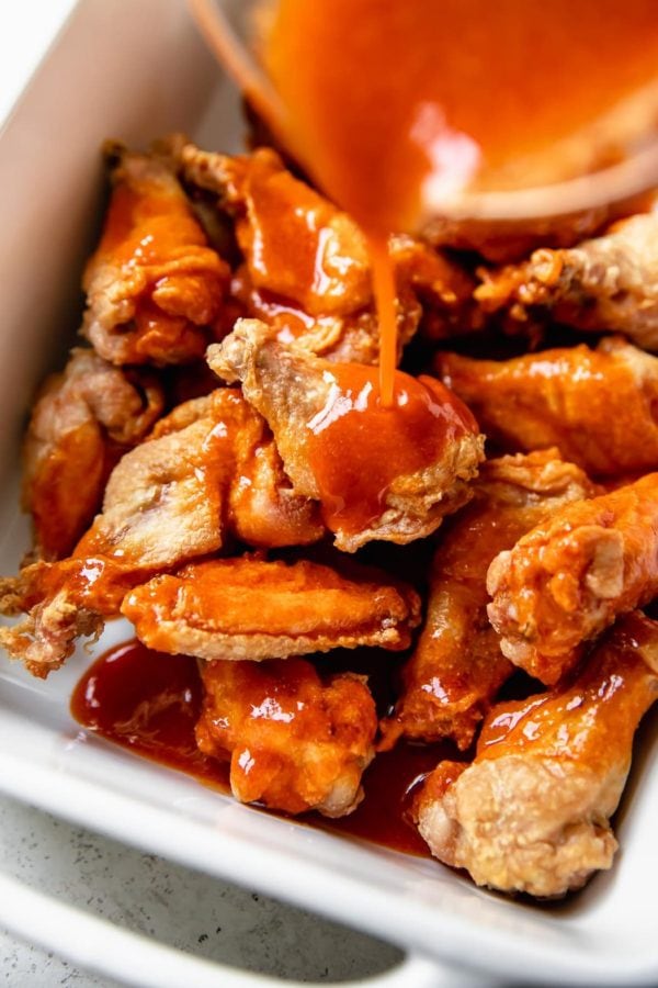 Crispy baked Buffalo wings in a white baking dish being doused in Buffalo sauce
