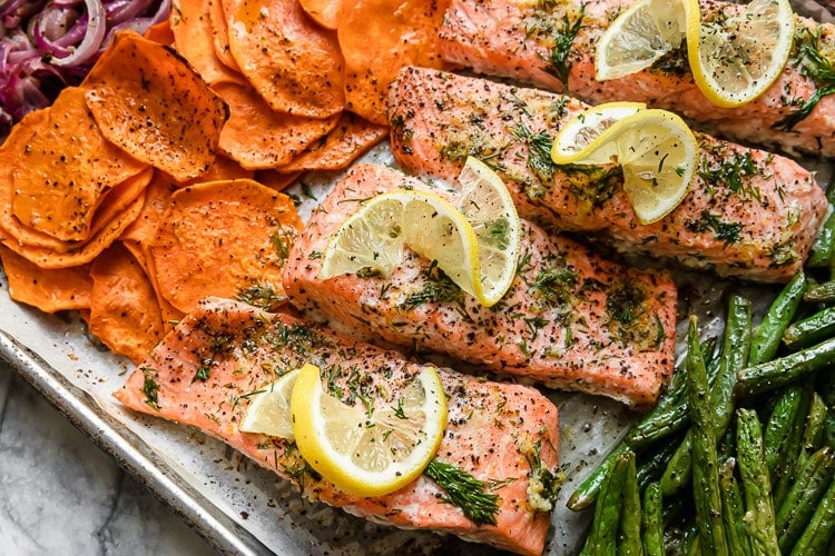 Overhead view baked salmon filets with seasoning on parchment lined baking sheet with sweet potato rounds.