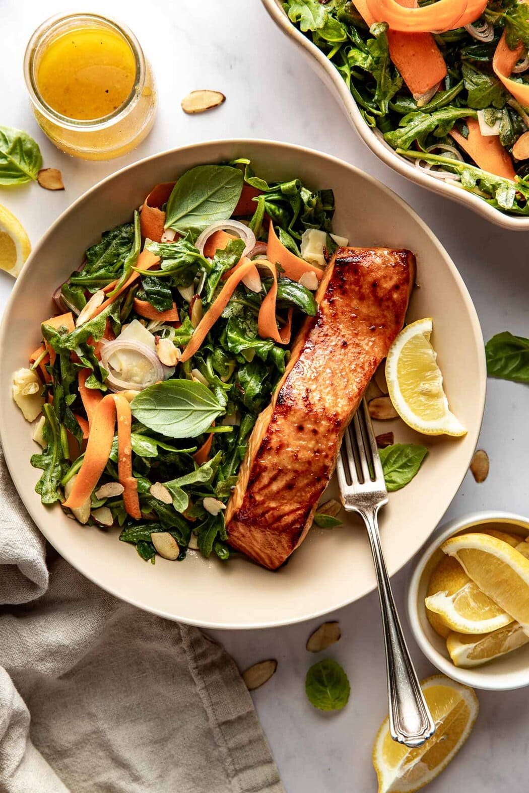 Close up view of a bowl of Arugula Salad with a piece of miso salmon on the side garnished with lemon wedges and fresh basil. 