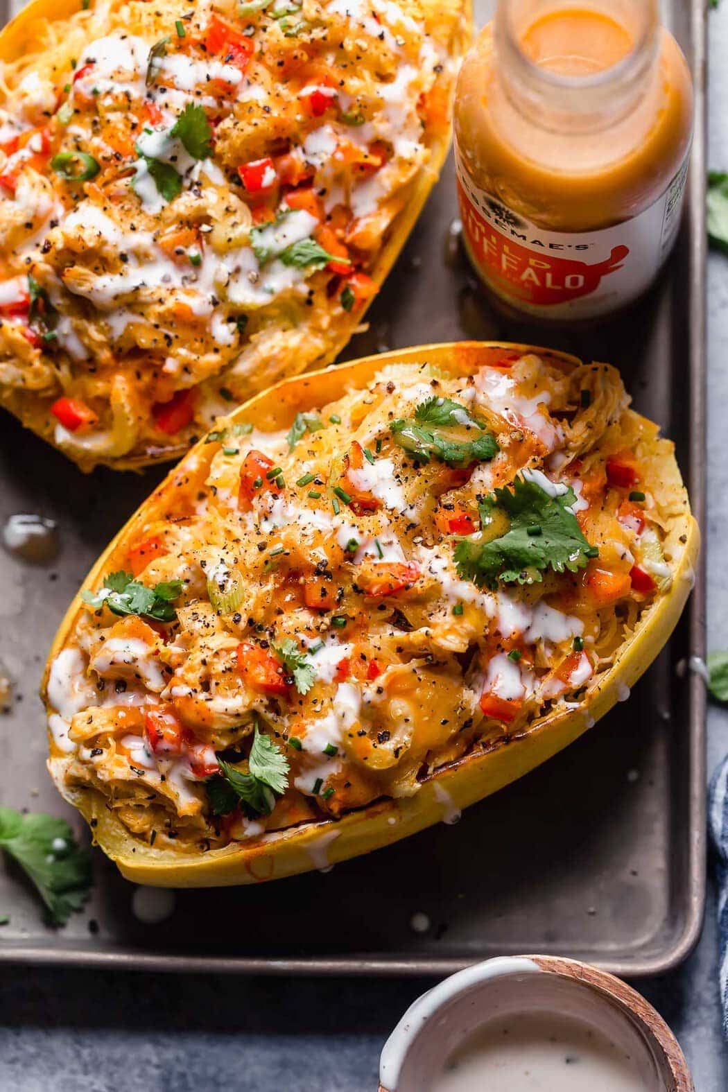 Buffalo chicken stuffed spaghetti squash halves on a baking sheet topped with fresh herbs, ranch dressing, and black pepper.