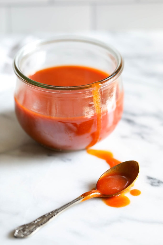 Vibrant orange/red homemade buffalo sauce in a short jar with a spoon laying in a puddle of sauce.
