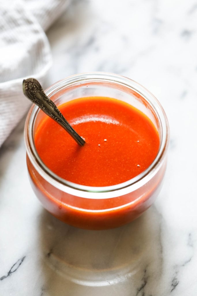 Homemade buffalo sauce in a short jar with a silver spoon in the spoon ready to stir