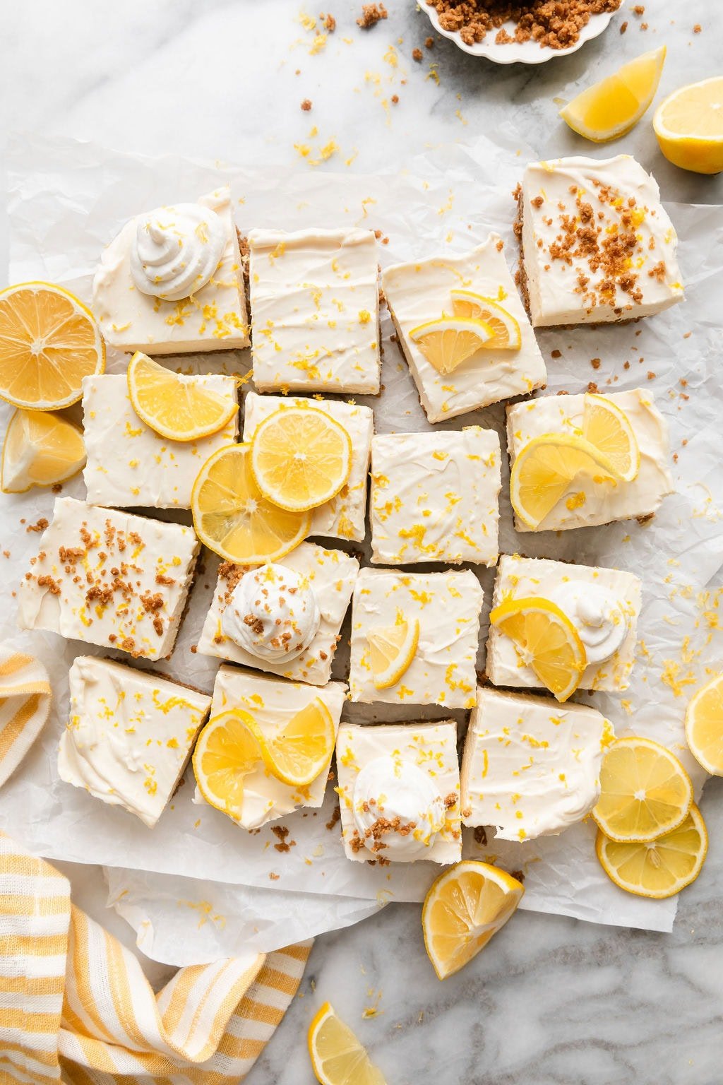 Overhead view of No Bake Lemon Cheesecake Bars cut into small squares on parchment paper topped with lemon zest and lemon slices. 