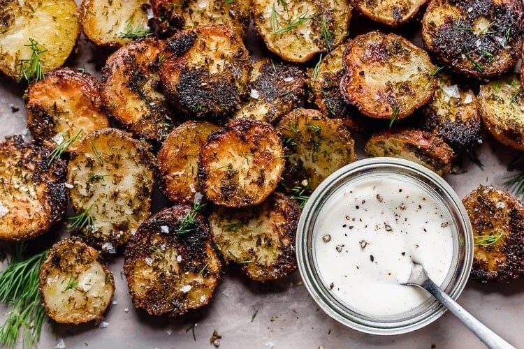 Crispy garlic ranch roasted potato halves on parchment sprinkled with dill weed and flaky sea salt, ranch dressing in a small bowl on the side.