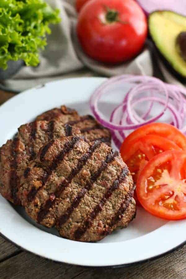 Grilled Bacon Burgers on a white plate with slice tomatoes and onion on the side