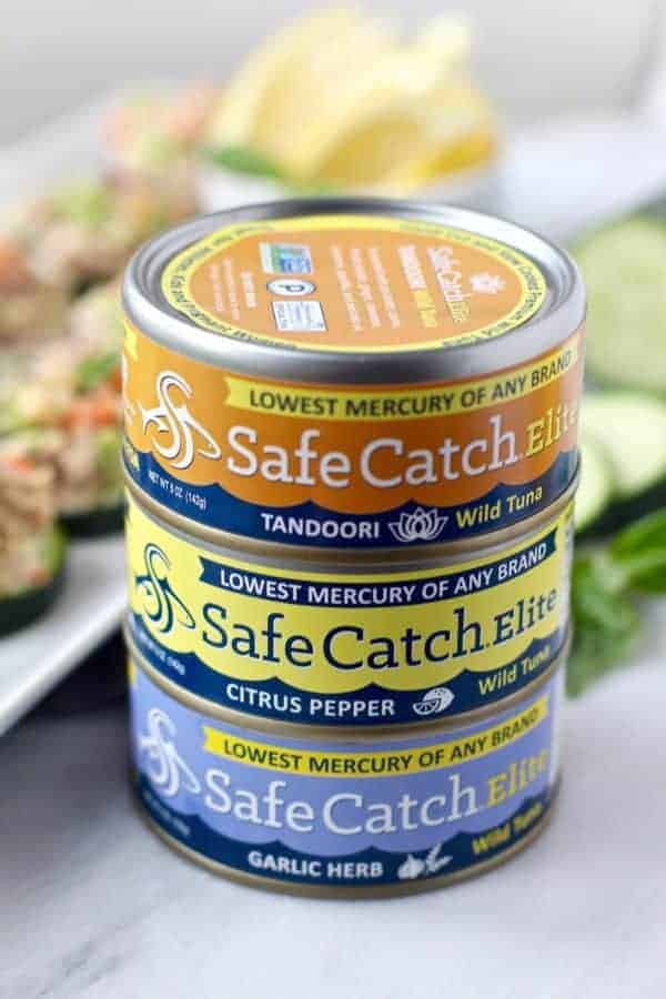 cans of SafeCatchElite Wild Tuna with a white plate of Cashew Tuna Salad Cucumber Bites in the background