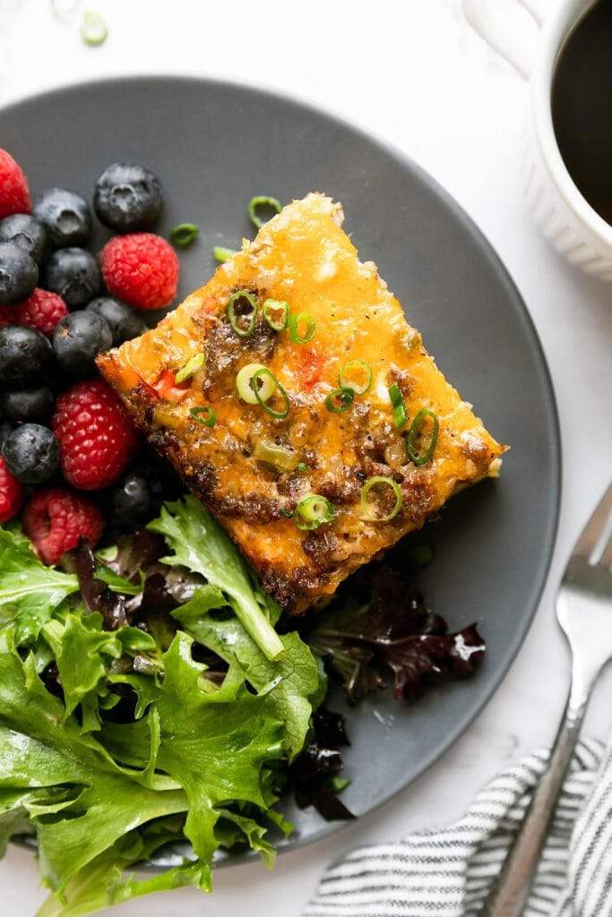 A piece of sausage egg bake on a dark grey plate with fresh berries and lettuce on the side. 