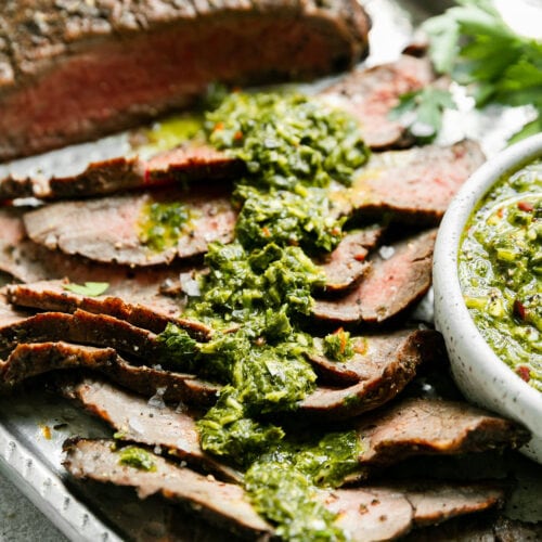 Close up side view thin slices of grilled flank steak with herby-green sauce spread over top.
