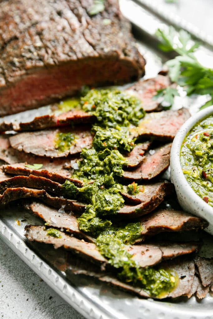 Close up side view thin slices of grilled flank steak with herby-green sauce spread over top. 