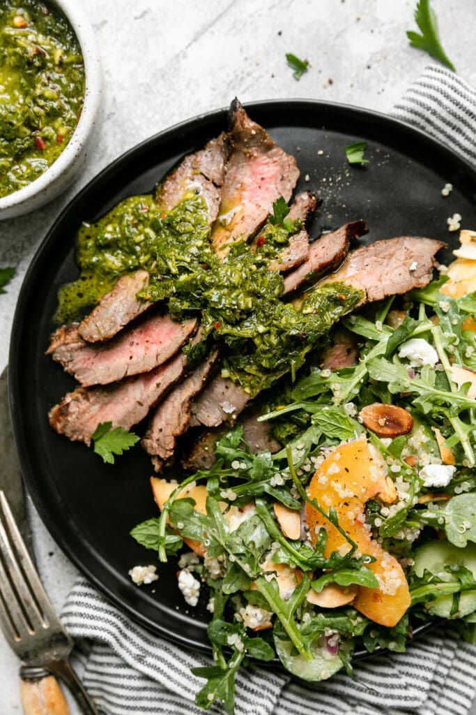 Overhead view black plate filled with serving of grilled flank steak with herby-green sauce over top and peach quinoa arugula salad on side. 