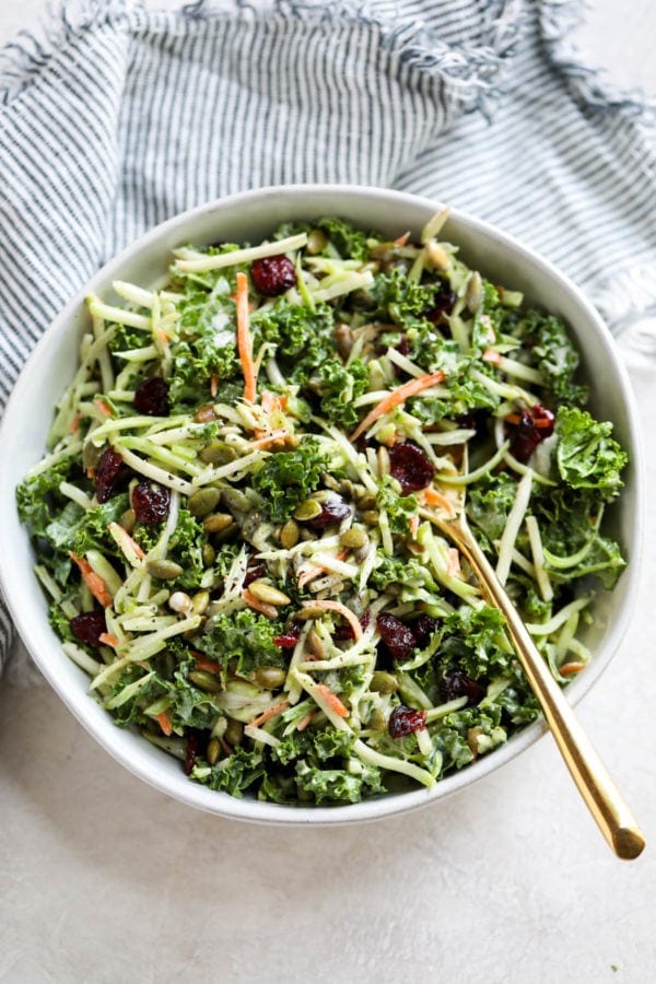 Birds eye view of creamy vegan broccoli slaw with pumpkin seeds and dried cranberries in a white bowl with a gold serving spoon.