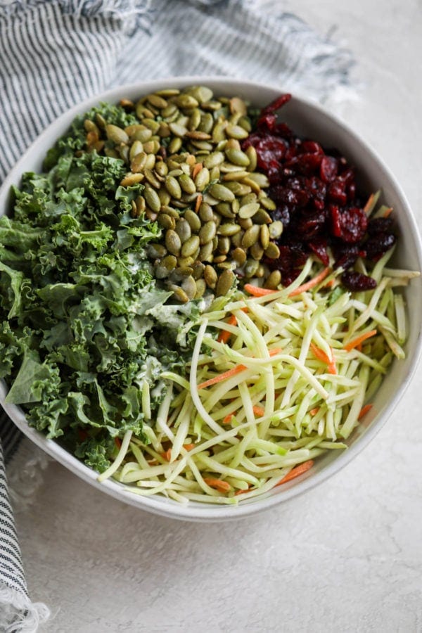 A white serving bowl filled with kale, pumpkin seeds, dried cranberries, and broccoli shreds.