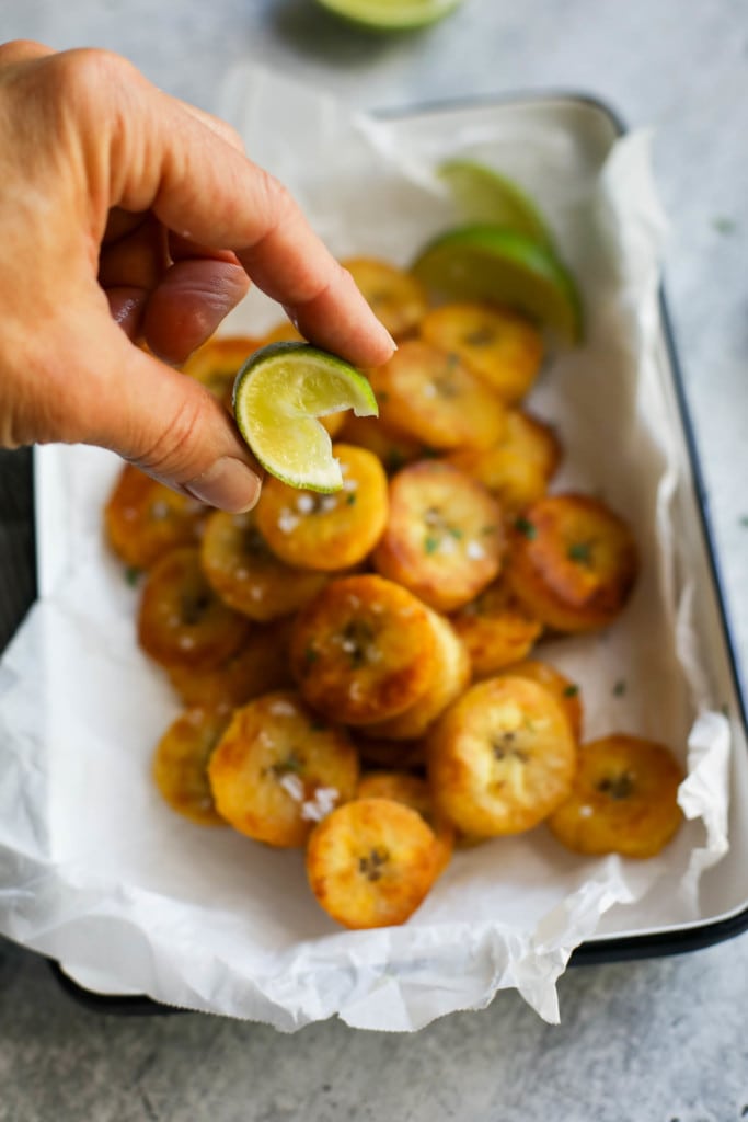A lime wedge being squeezed over a pile of freshly fried plantain rounds. 