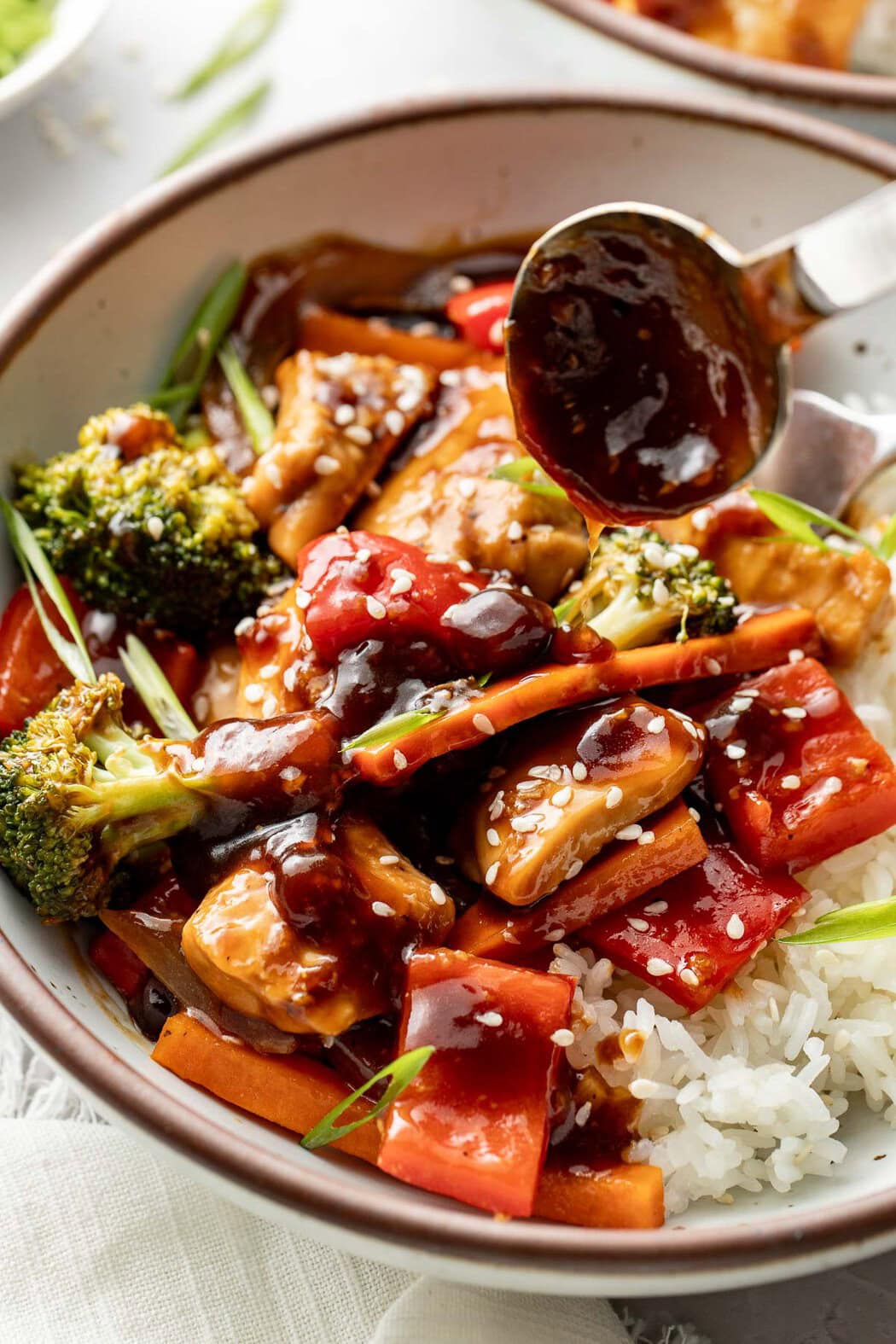 A spoon drizzling teriyaki sauce over a bowl of rice topped with sheet pan teriyaki chicken and vegetables.