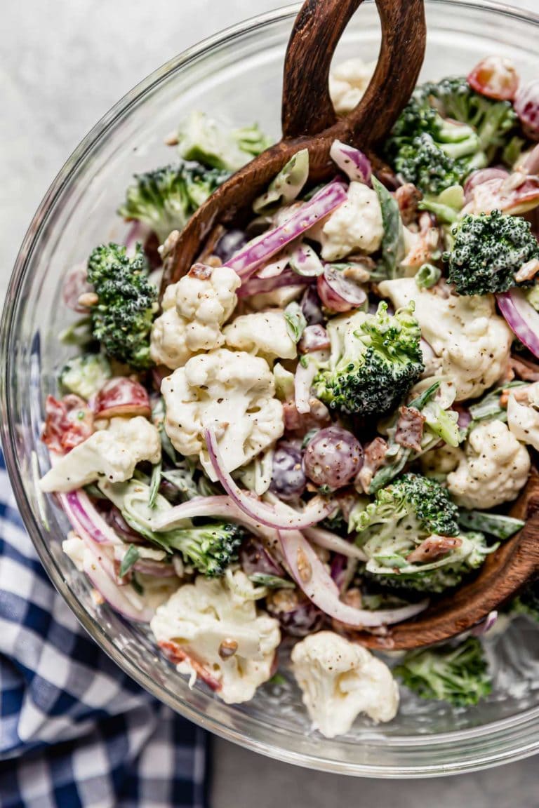 Broccoli cauliflower salad in a clear glass bowl with two wooden spoons for serving. 