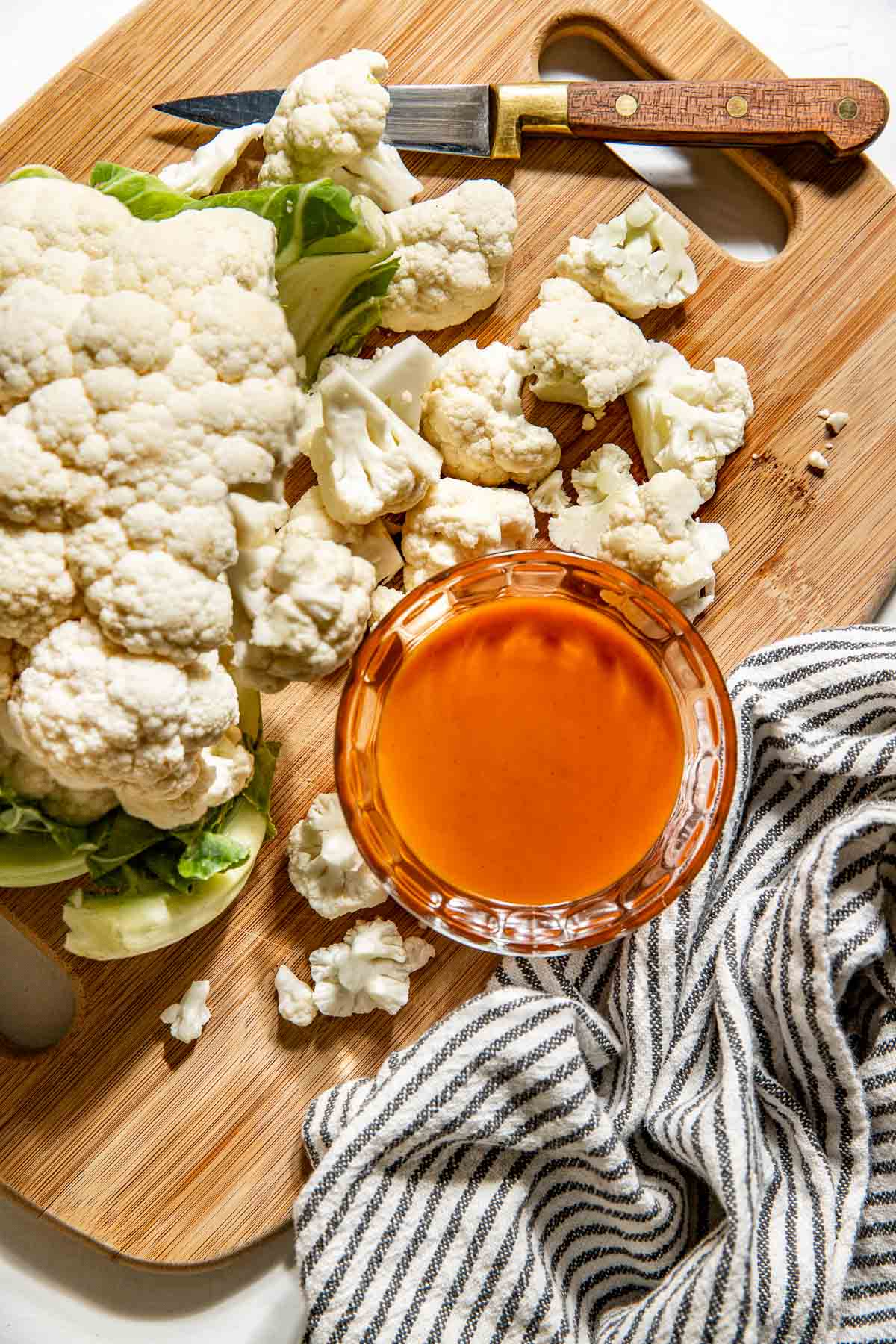 Overhead view of a head of cauliflower being cut into pieces on a cutting board next to buffalo sauce. 