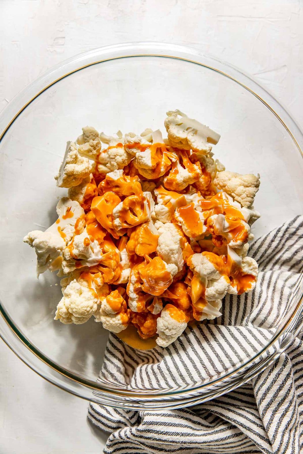 Overhead view of cauliflower pieces in a glass bowl topped with buffalo sauce ready for tossing. 