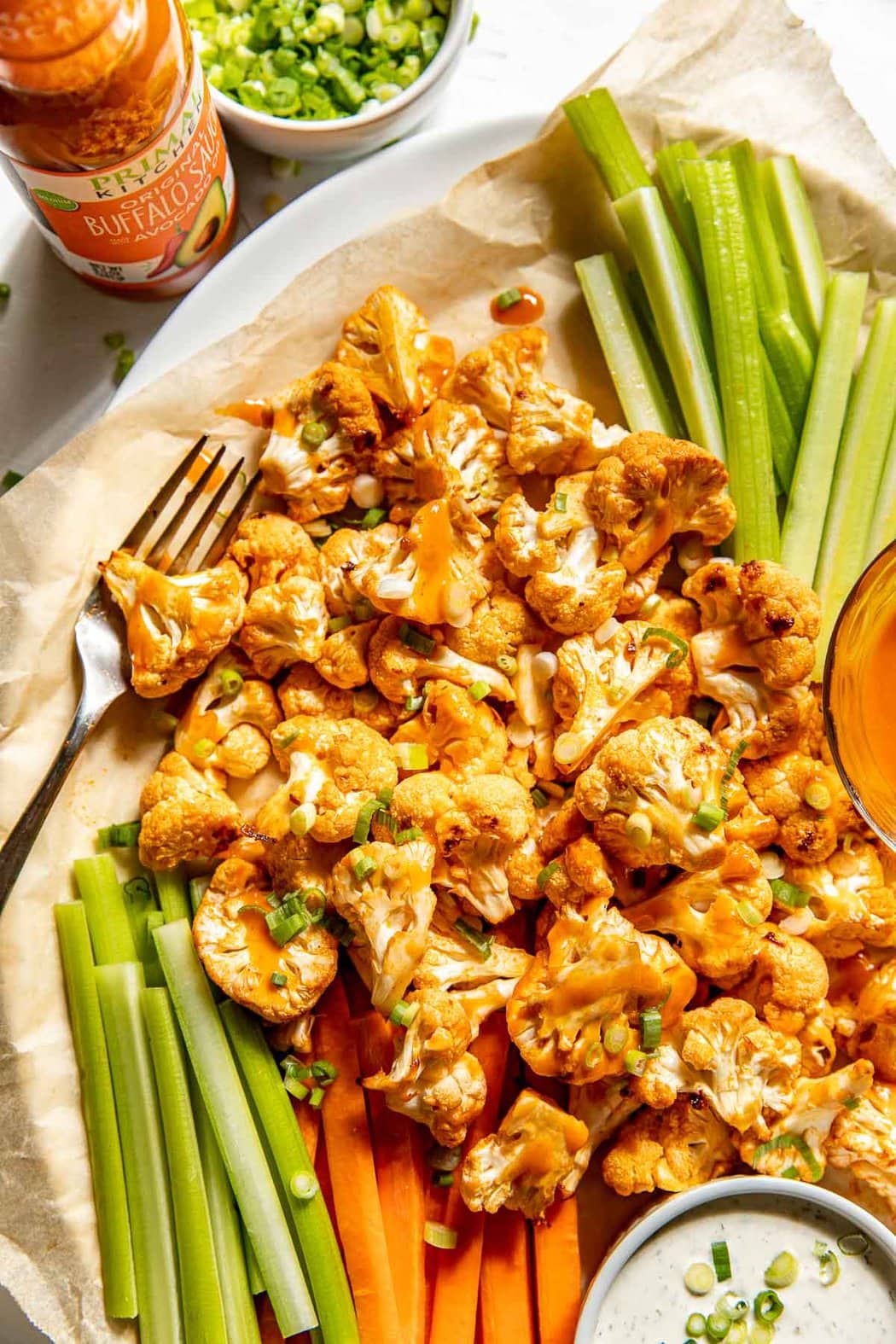 Close up view of a sheet pan of freshly roasted Buffalo Cauliflower Bites next to celery sticks and buffalo sauce, topped with green onions. 