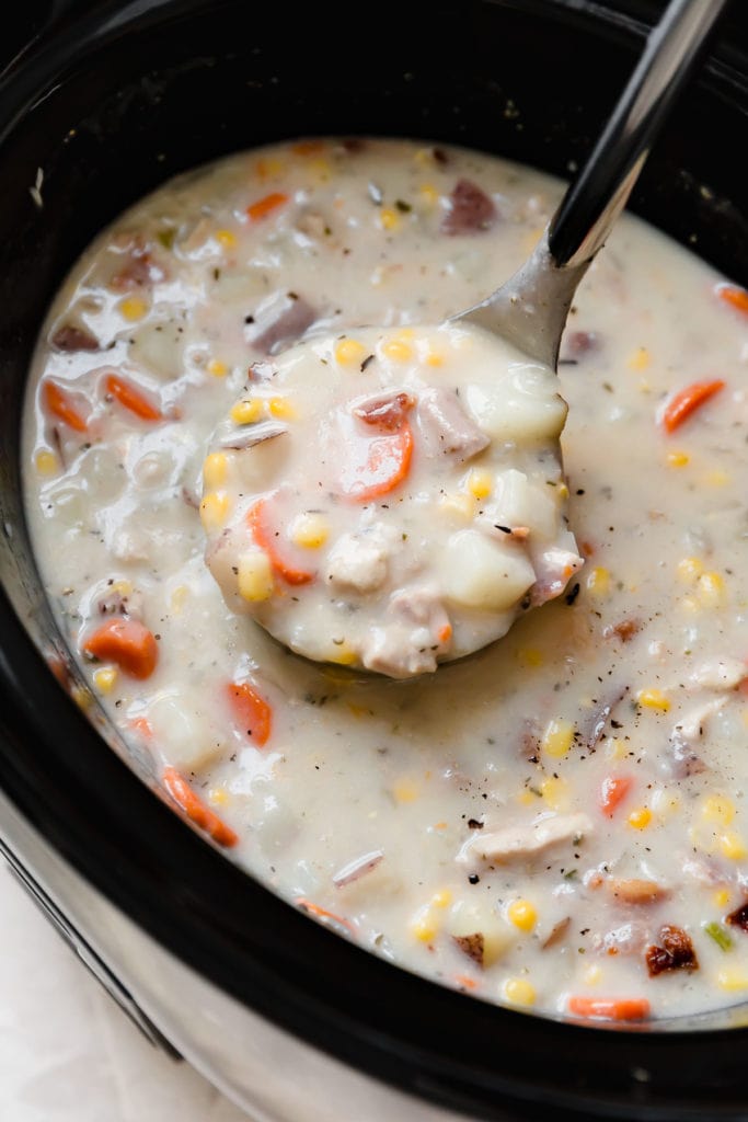 A ladle full of creamy slow cooker chicken corn chowder filled with large chunks of carrot, potato, and corn kernels.