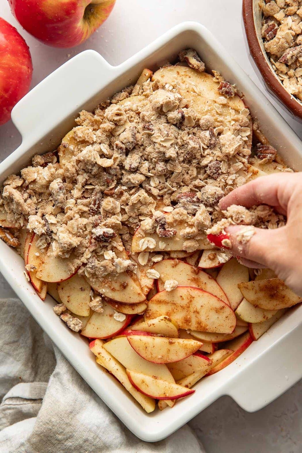 Crumble topping being sprinkled over sliced apples in a square baking dish.