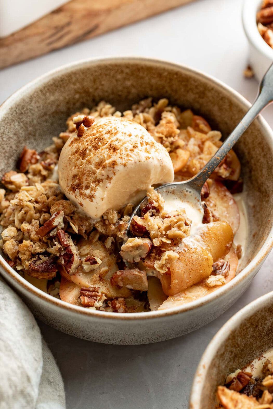 A bowl of healthy apple crisp topped with vanilla ice cream, a spoon holding a bite on top.