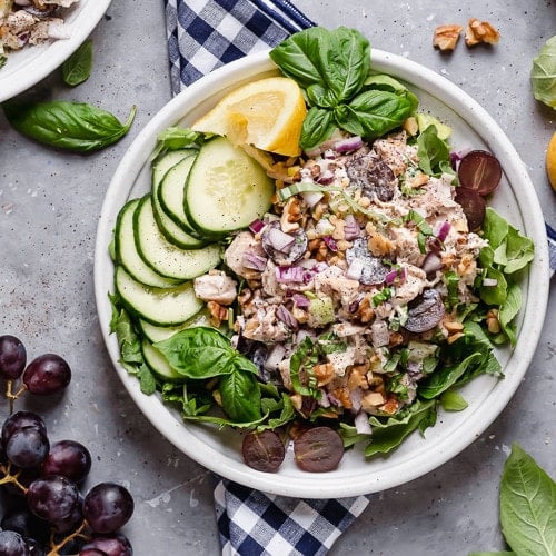 Overhead view of creamy Waldorf chicken salad on a white plate over a bed of greens with grapes and cucumbers on the side.